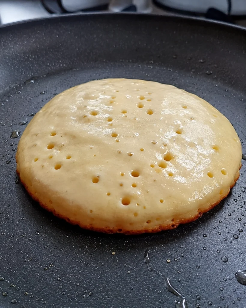 Pancake cocinándose en sartén antiadherente con burbujas visibles en la superficie.