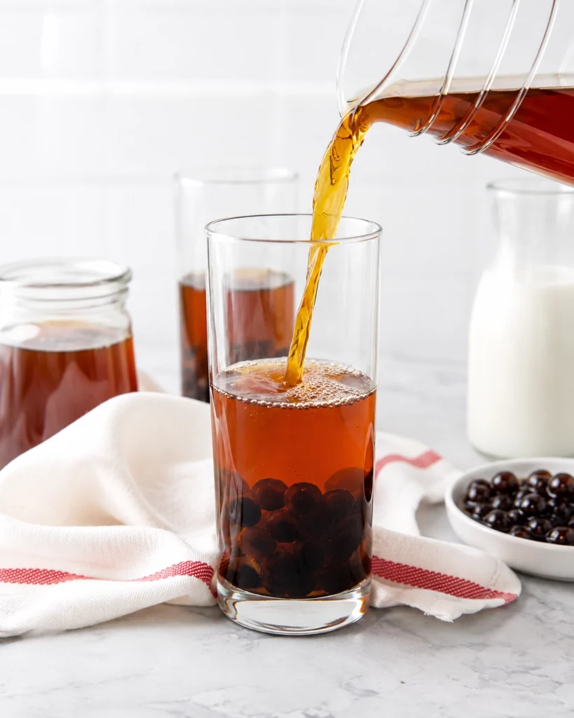 Té negro ámbar vertiéndose desde una jarra de vidrio en un vaso con perlas de tapioca en el fondo.