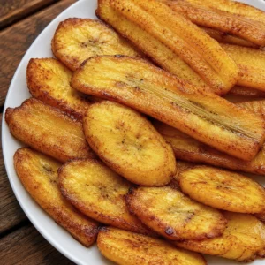 Plátanos maduros fritos 7 Plátanos maduros fritos dorados y caramelizados servidos en un plato blanco sobre una mesa de madera.