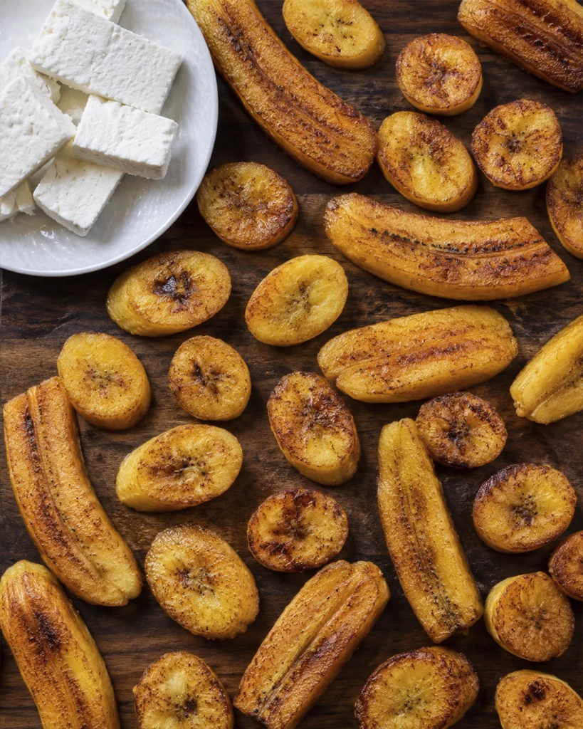 Plátanos maduros fritos 6 Plátanos maduros fritos en tajadas y mitades, acompañados con queso fresco sobre una tabla de madera.