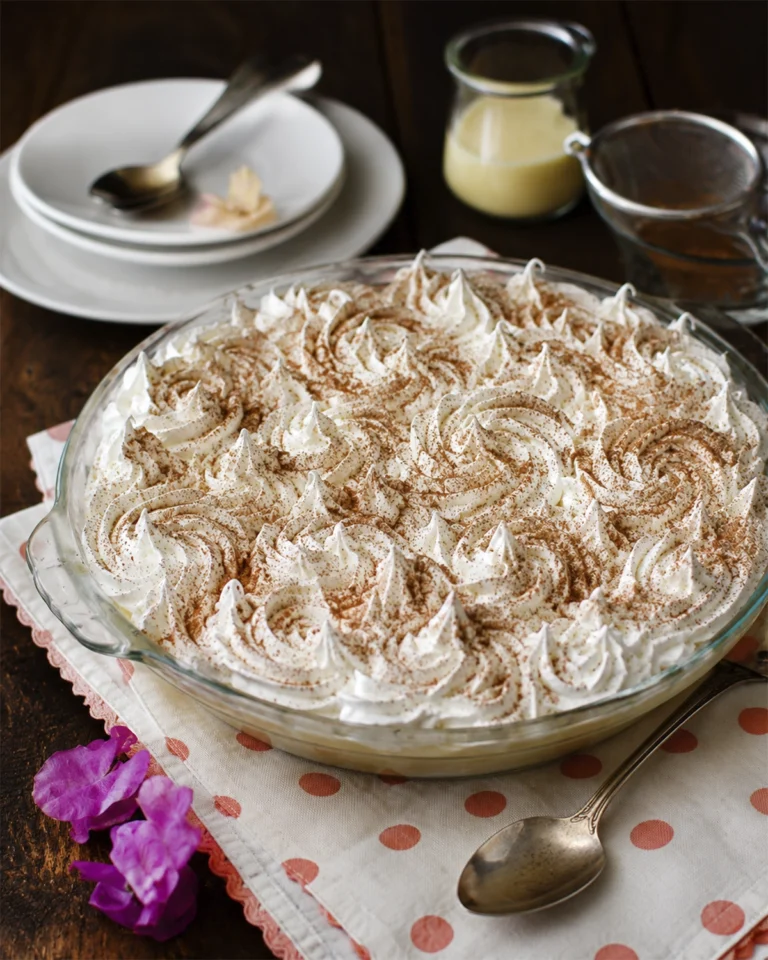 Torta tres leches con merengue blanco y canela en un molde de vidrio sobre mesa de madera.