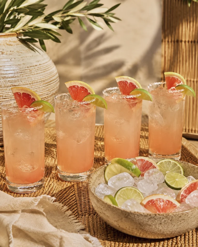 Cuatro vasos de coctel Paloma con tequila con hielo, toronja, lima y borde de sal sobre una mesa.